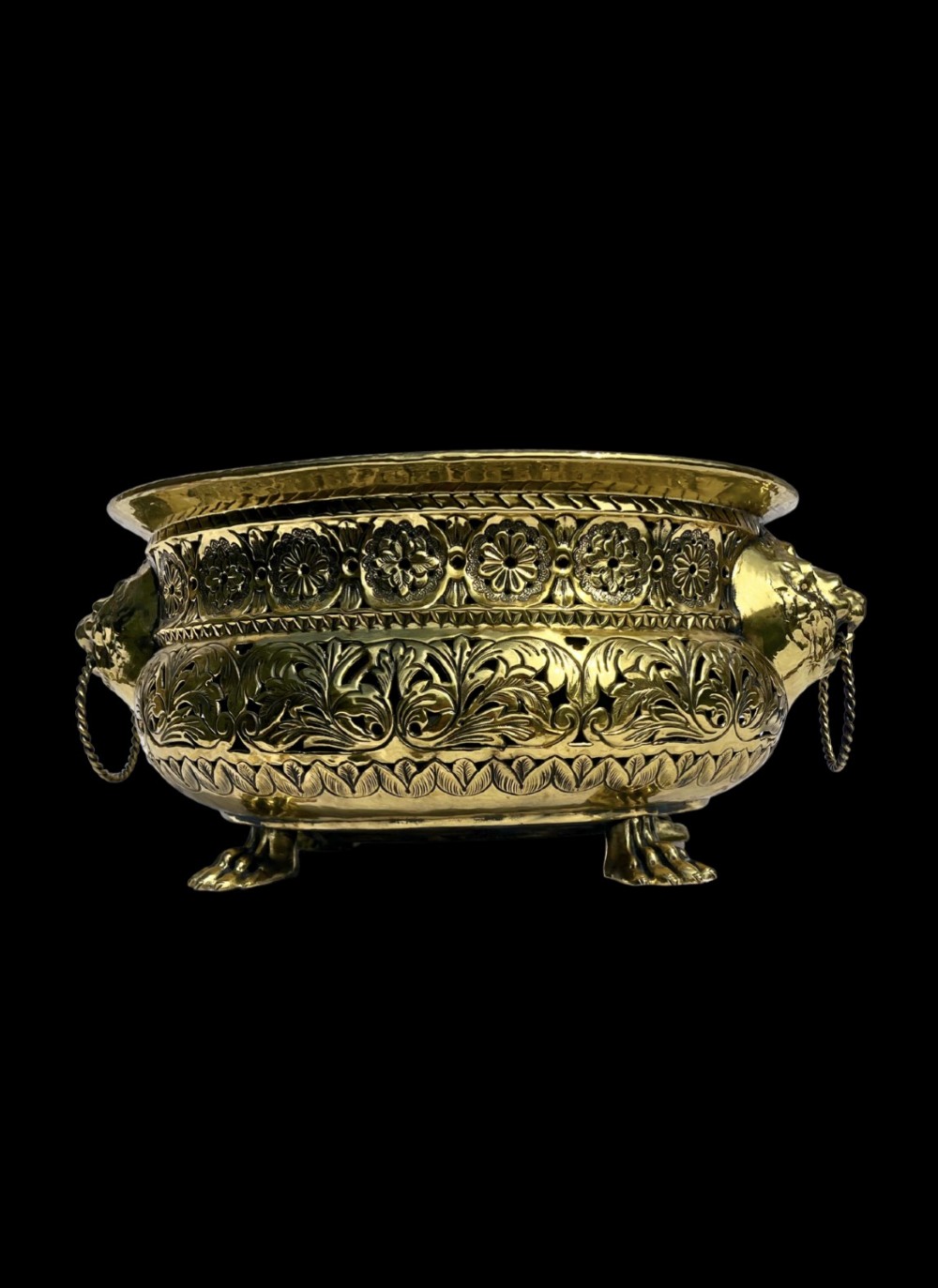 A round antique Dutch brass jardiniere featuring intricate repousse foliate patterning, dual lion-head ring handles, and claw feet.