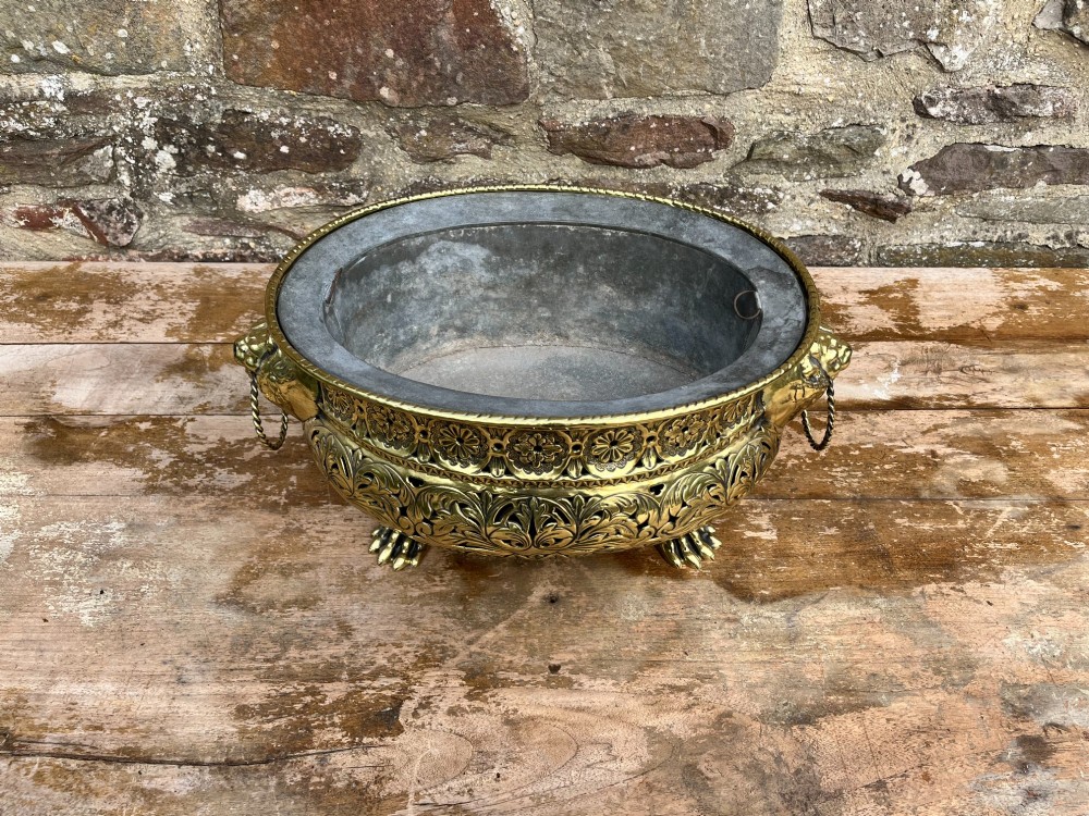 A round antique Dutch brass jardiniere featuring intricate repousse foliate patterning, dual lion-head ring handles, and claw feet. View IV