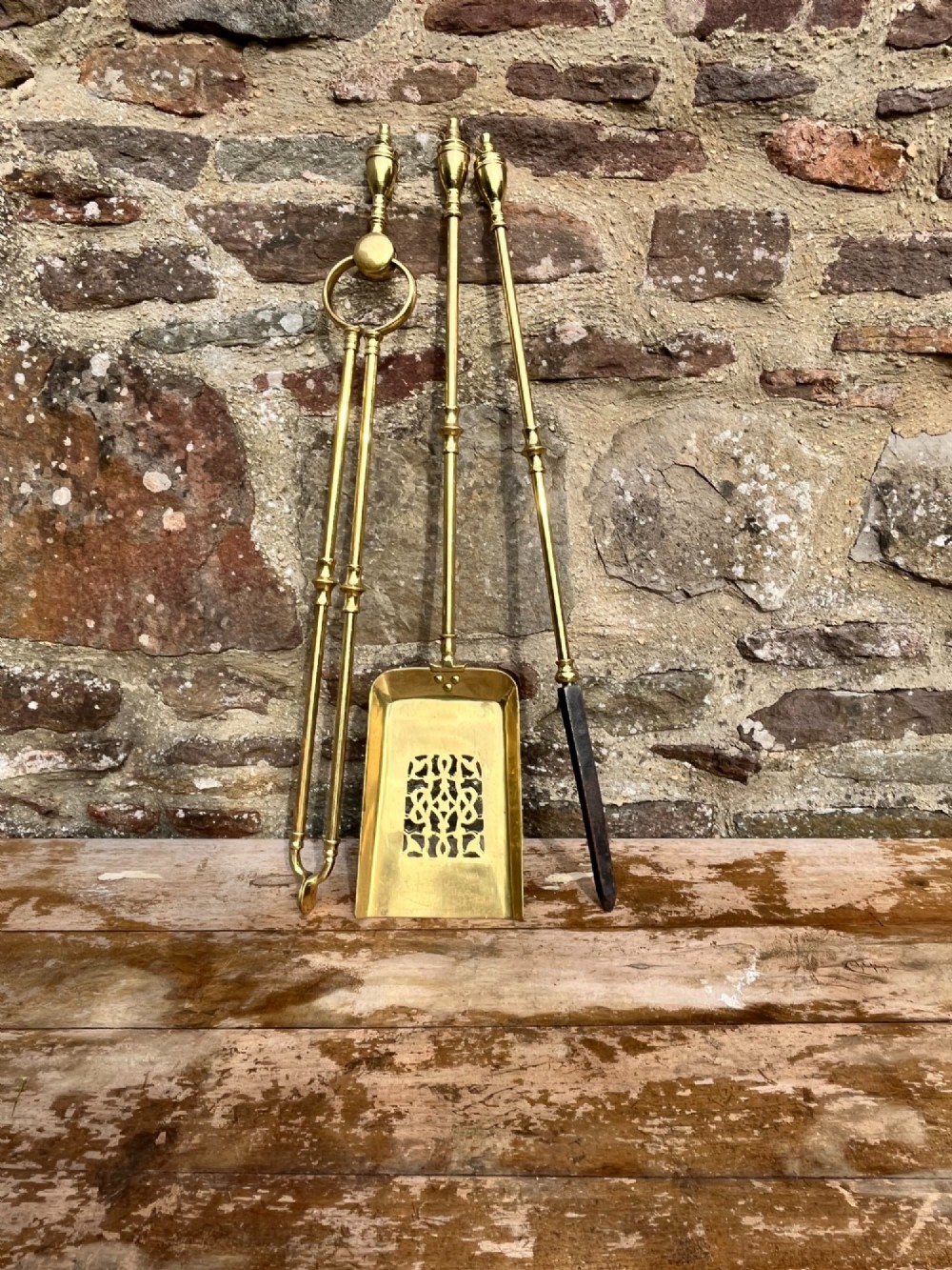 A three-piece set of polished brass fire tools featuring a pierced tray shovel, a poker, and tongs, all decorated with urn-shaped finials. View III