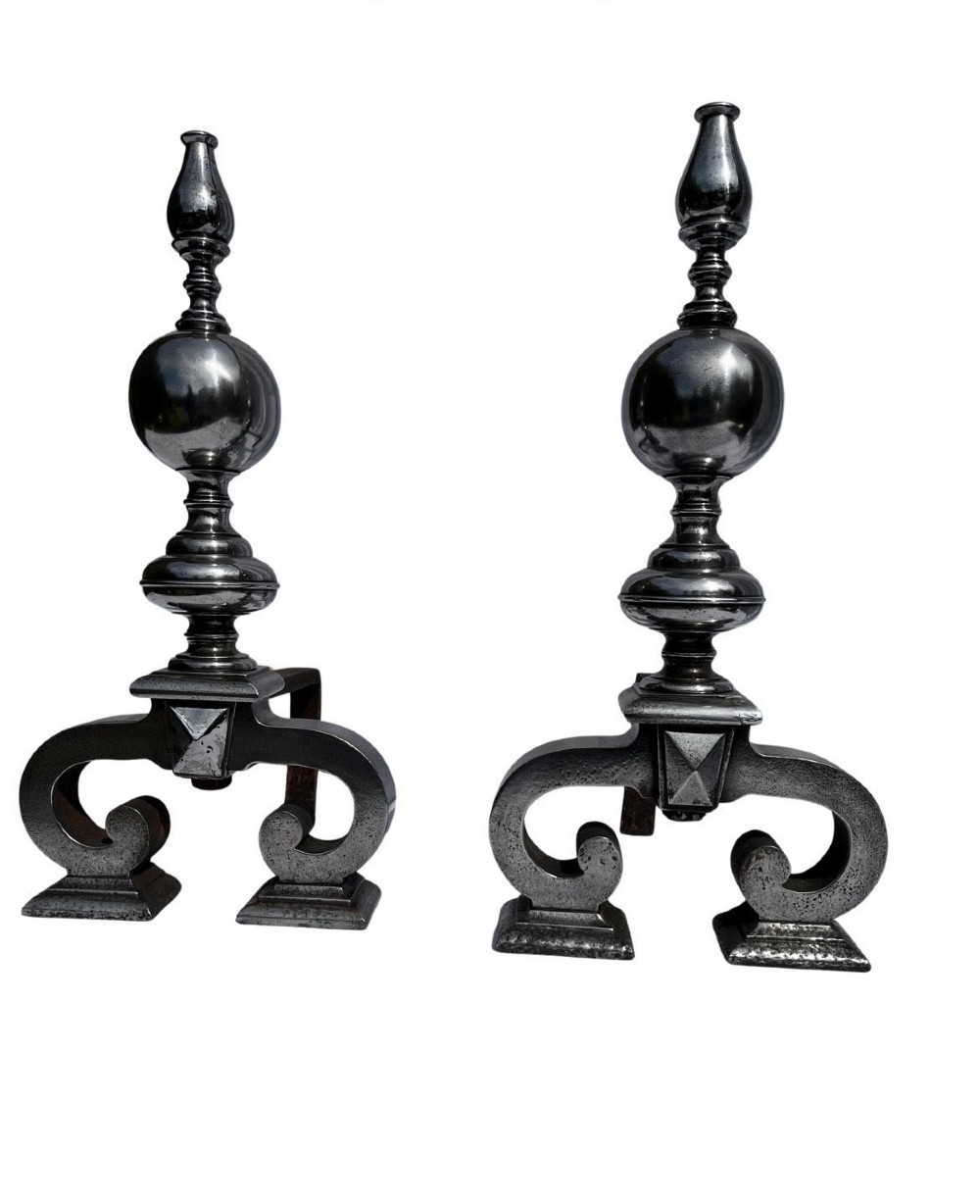 A pair of polished iron fire dogs featuring turned spherical finials and scrolled feet.
