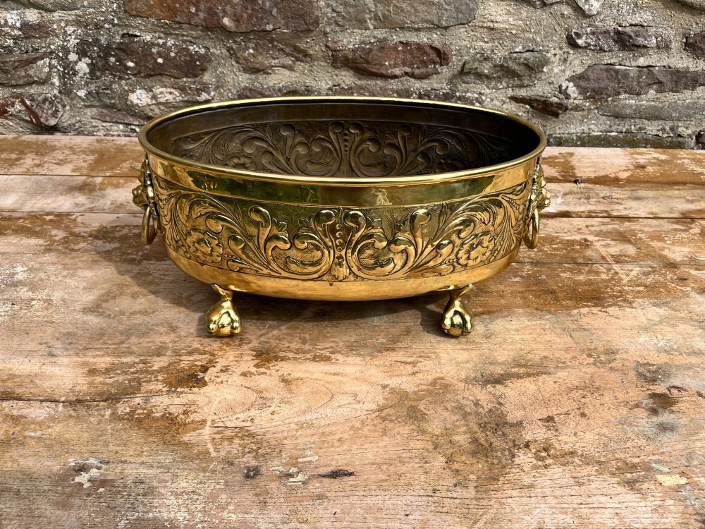 An oval brass jardiniere featuring embossed floral scrollwork, lion mask ring handles, and four claw feet. View IV