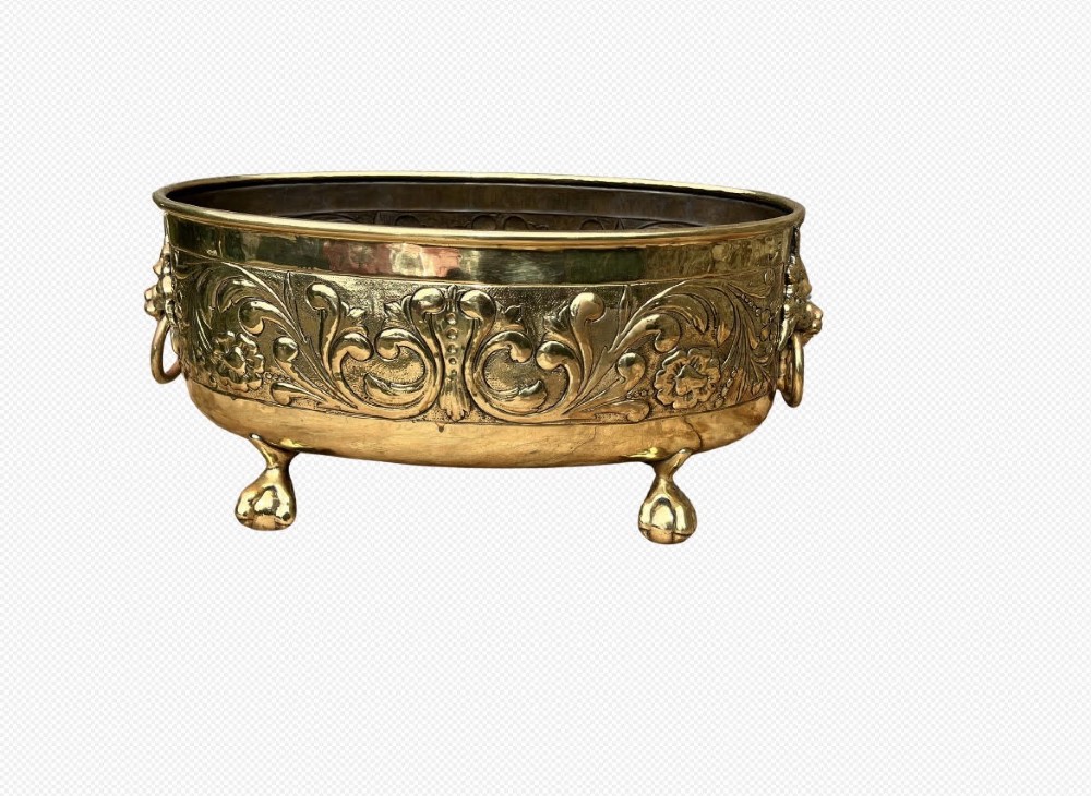 An oval brass jardiniere featuring embossed floral scrollwork, lion mask ring handles, and four claw feet.