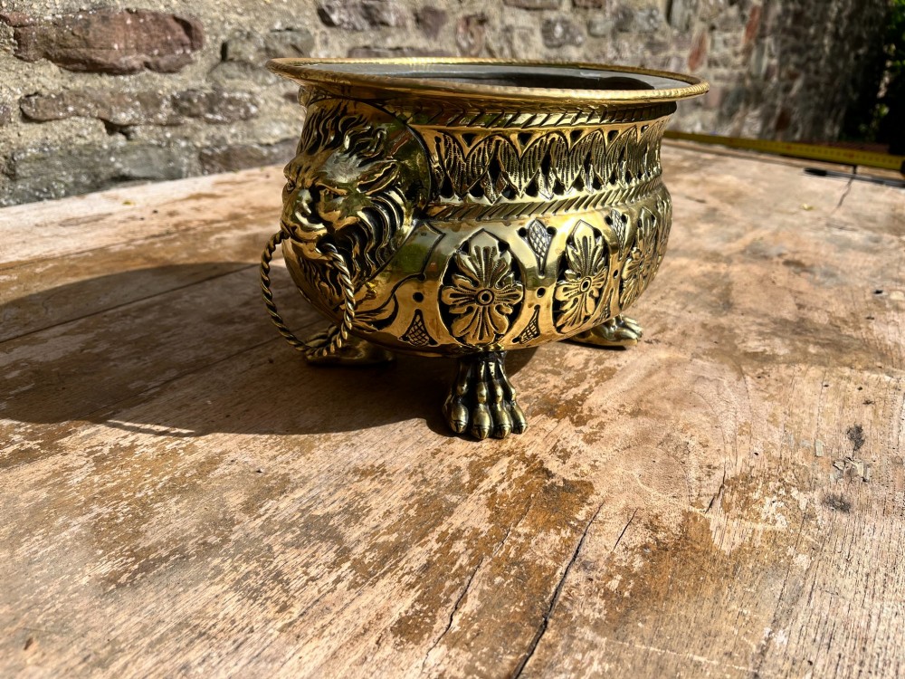 An antique oval pierced brass jardiniere featuring floral repousse panels, lion mask handles with drop rings, and claw feet. View IV