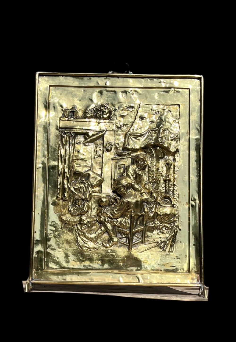 An antique rectangular brass fire screen featuring a detailed repousse embossed genre scene of figures in a room. View II
