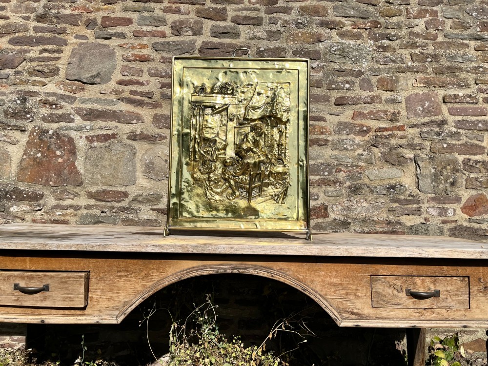 An antique rectangular brass fire screen featuring a detailed repousse embossed genre scene of figures in a room. View III