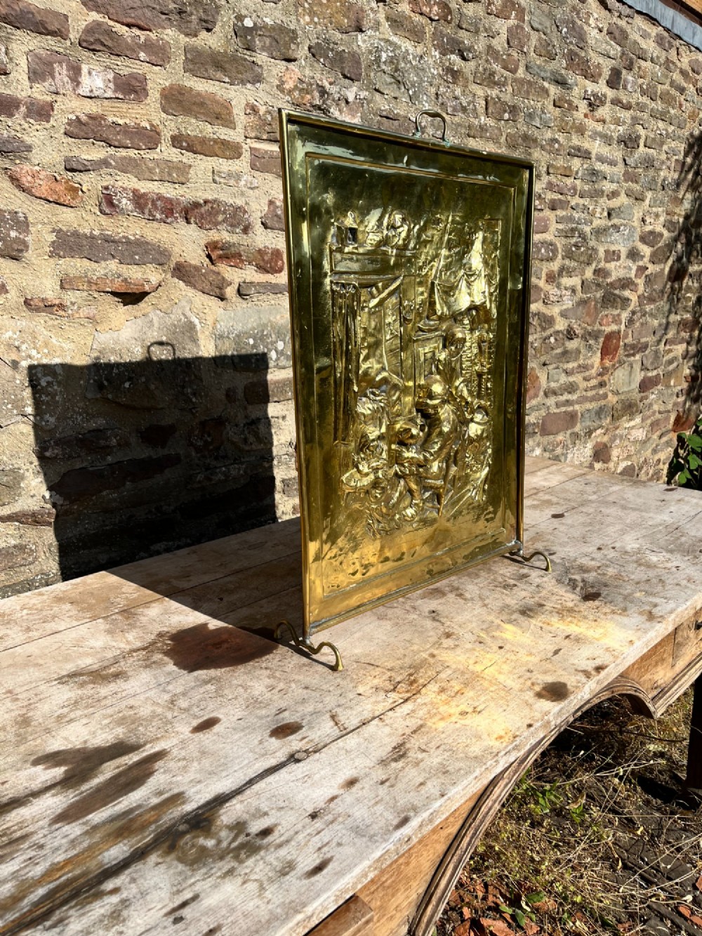 An antique rectangular brass fire screen featuring a detailed repousse embossed genre scene of figures in a room. View IV