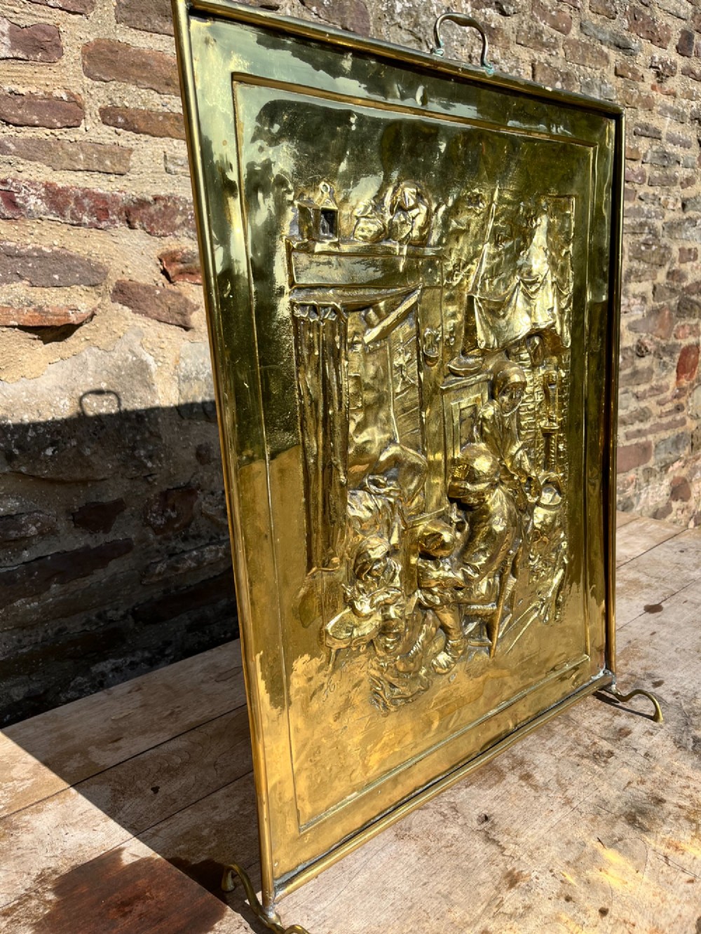 An antique rectangular brass fire screen featuring a detailed repousse embossed genre scene of figures in a room. View V