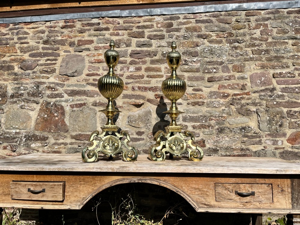 A pair of decorative polished brass French fire dogs with urn finials, gadrooned details, scrolled bases, and central fleur-de-lis motifs. View III