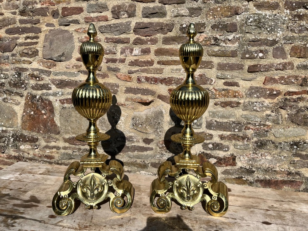 A pair of decorative polished brass French fire dogs with urn finials, gadrooned details, scrolled bases, and central fleur-de-lis motifs. View IV