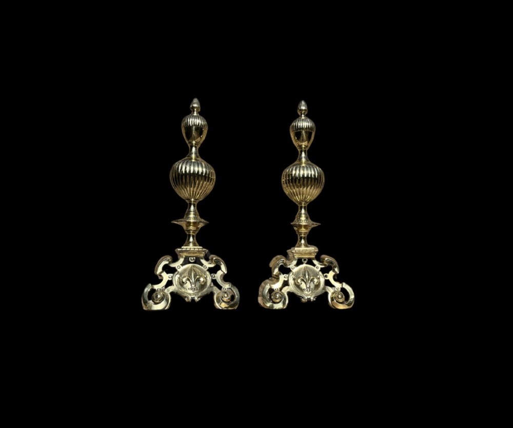 A pair of decorative polished brass French fire dogs with urn finials, gadrooned details, scrolled bases, and central fleur-de-lis motifs. View V