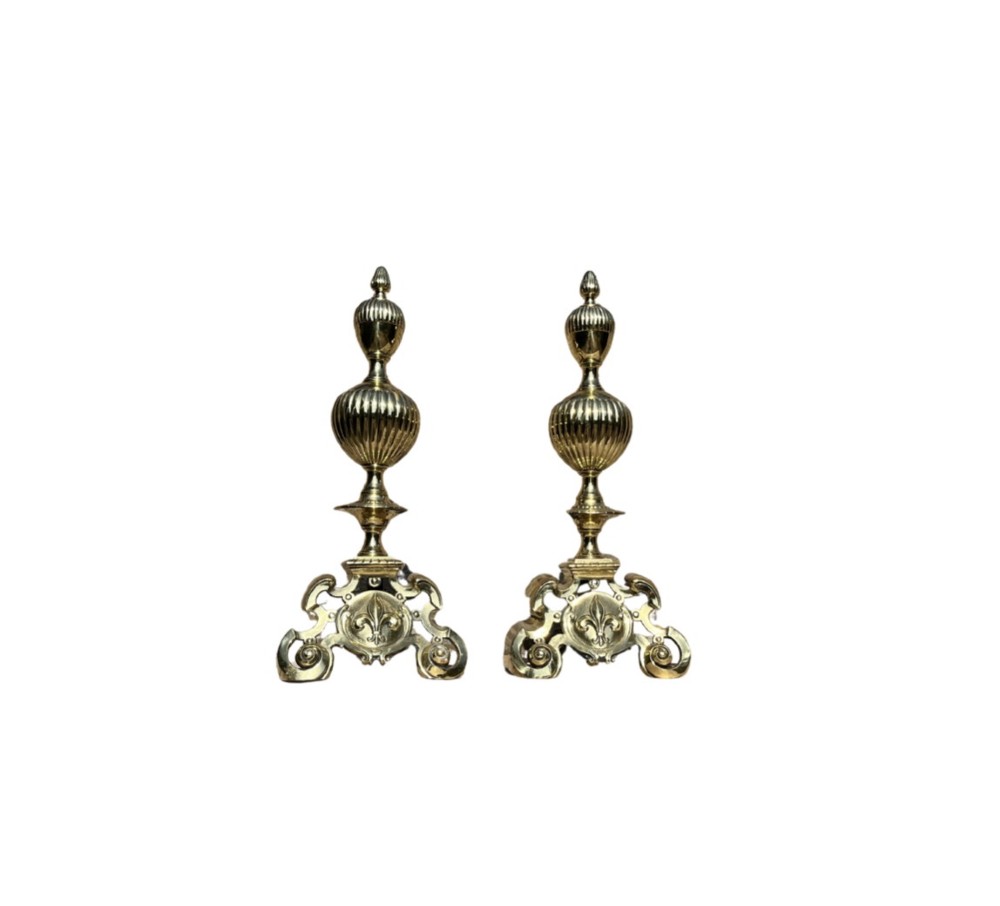 A pair of decorative polished brass French fire dogs with urn finials, gadrooned details, scrolled bases, and central fleur-de-lis motifs.