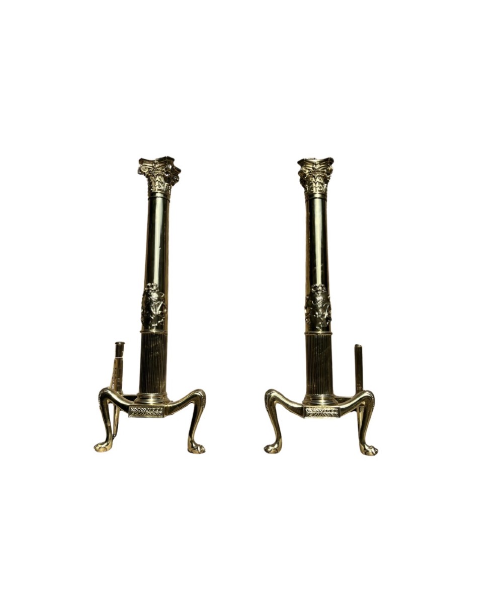 A pair of tall polished brass fire dogs featuring fluted Corinthian columns with relief crests and cabriole legs ending in claw feet.
