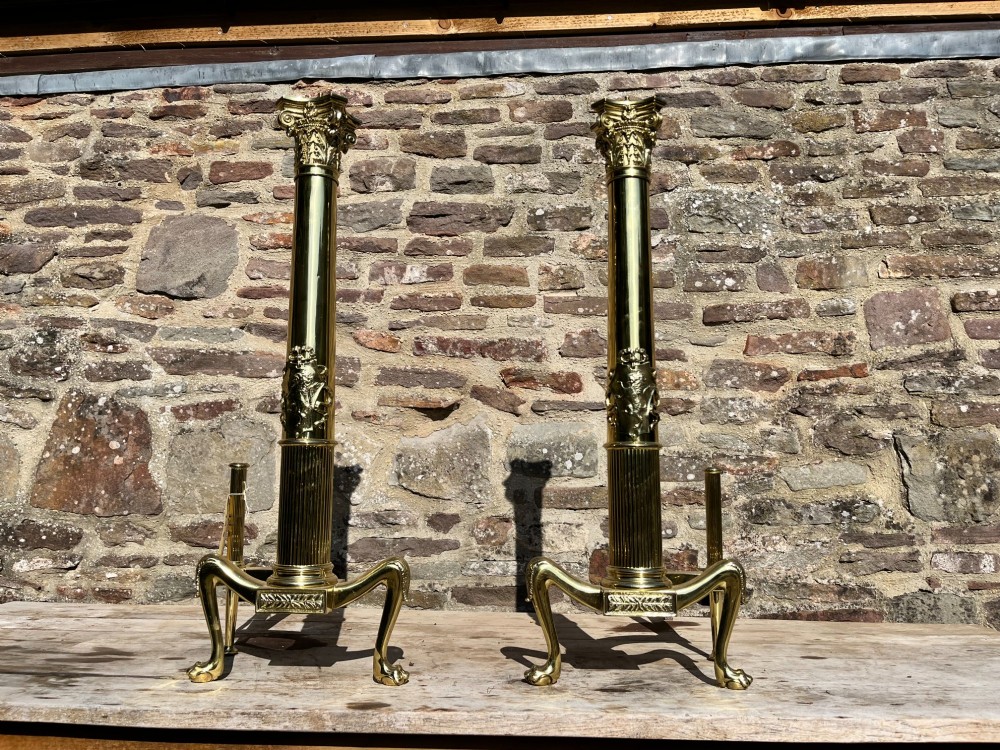 A pair of tall polished brass fire dogs featuring fluted Corinthian columns with relief crests and cabriole legs ending in claw feet. View VII