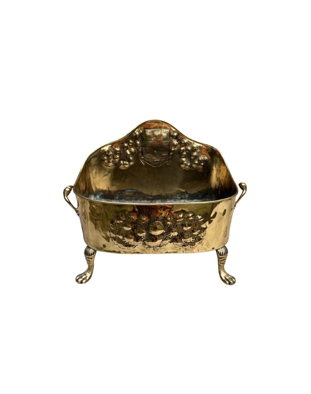 An antique brass hearth tidy featuring embossed floral crest decoration, twin side handles, and cast paw feet.
