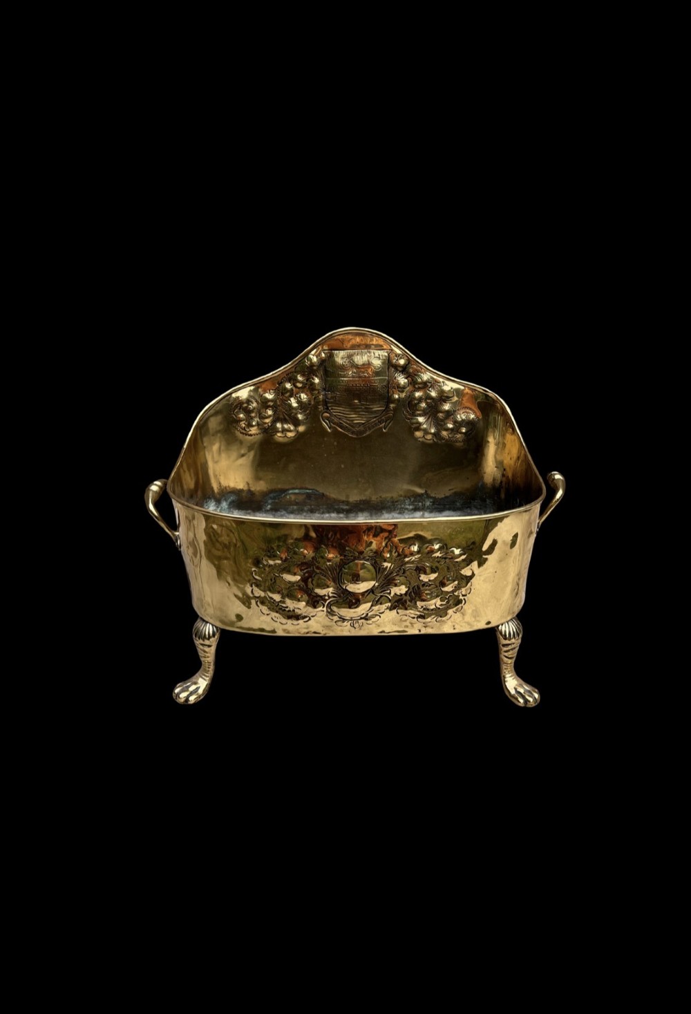 An antique brass hearth tidy featuring embossed floral crest decoration, twin side handles, and cast paw feet. View II