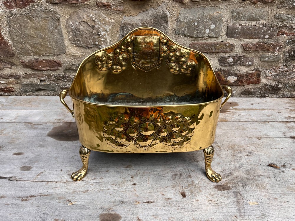 An antique brass hearth tidy featuring embossed floral crest decoration, twin side handles, and cast paw feet. View III