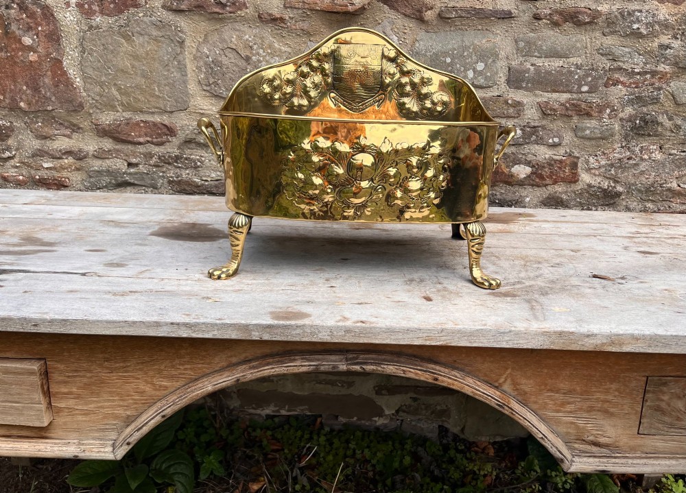 An antique brass hearth tidy featuring embossed floral crest decoration, twin side handles, and cast paw feet. View IV