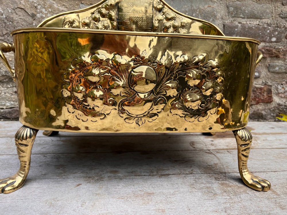 An antique brass hearth tidy featuring embossed floral crest decoration, twin side handles, and cast paw feet. View V