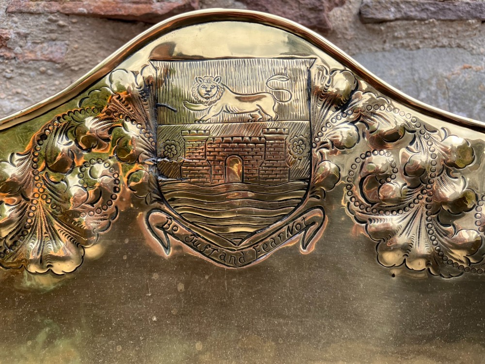 An antique brass hearth tidy featuring embossed floral crest decoration, twin side handles, and cast paw feet. View VI