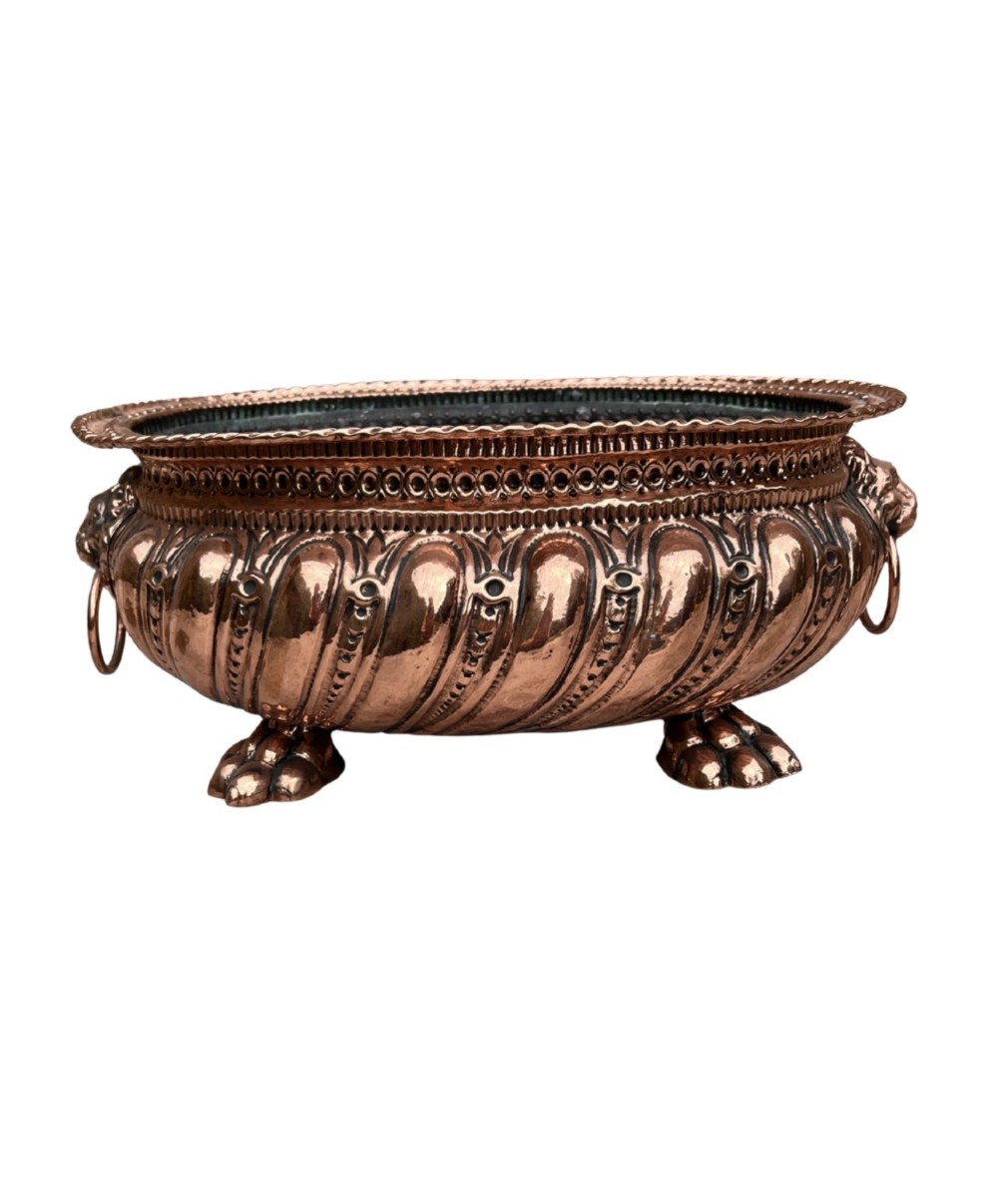 An oval antique copper jardiniere featuring gadrooned body panels, lion mask drop-ring handles, and cast paw feet.