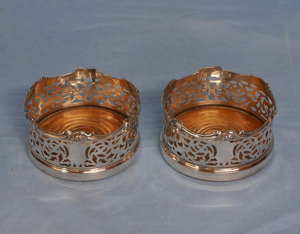 A pair of circular silver-plated wine coasters featuring pierced scrollwork sides and turned wooden bases.