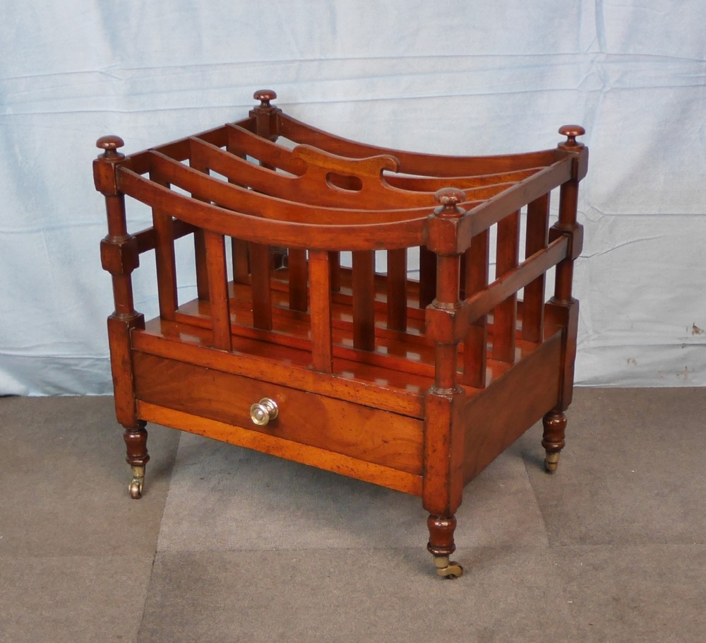 An antique mahogany canterbury with four slatted compartments, a curved central handle, turned posts, a single lower drawer, and brass castors.