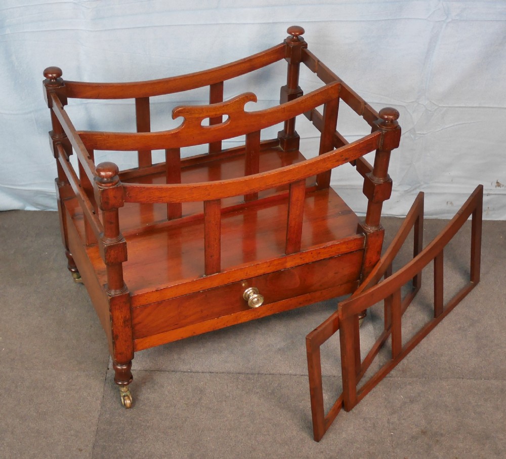 An antique mahogany canterbury with four slatted compartments, a curved central handle, turned posts, a single lower drawer, and brass castors. View IV