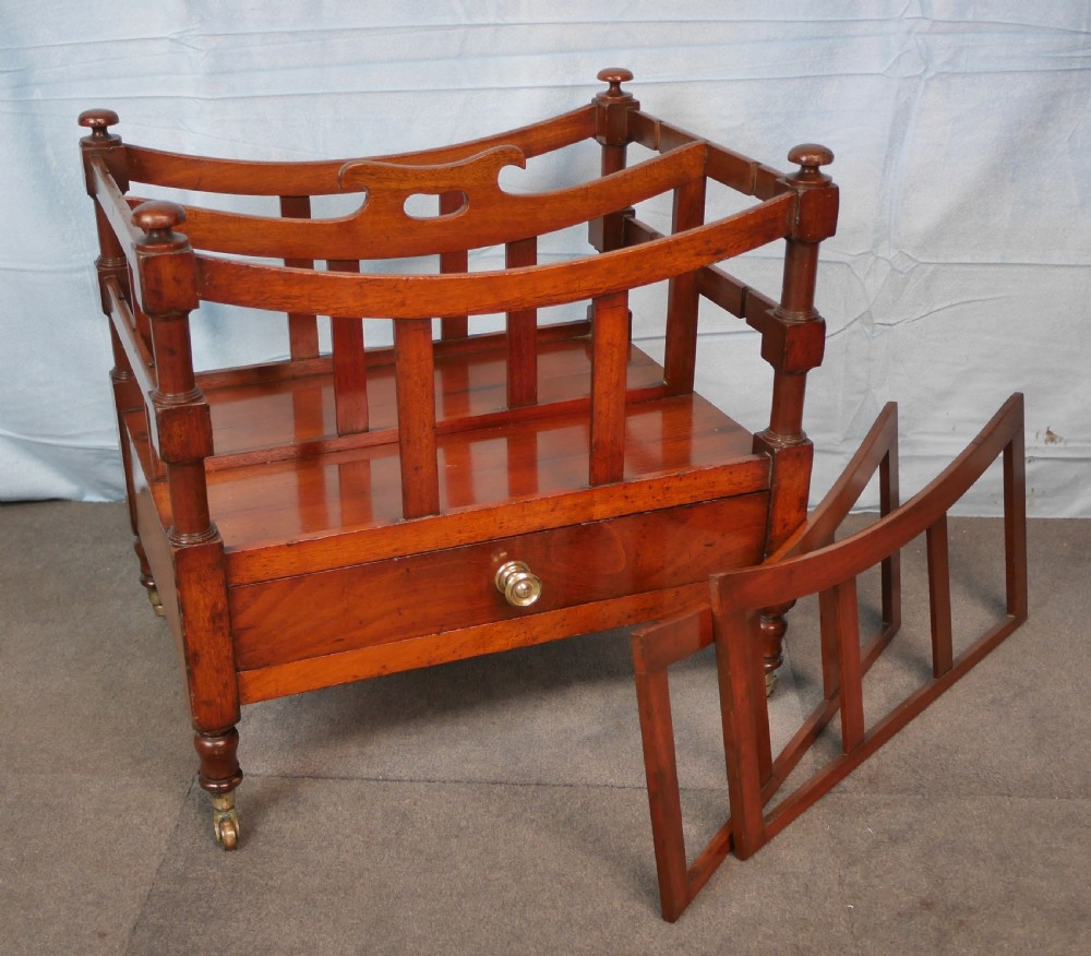 An antique mahogany canterbury with four slatted compartments, a curved central handle, turned posts, a single lower drawer, and brass castors. View V