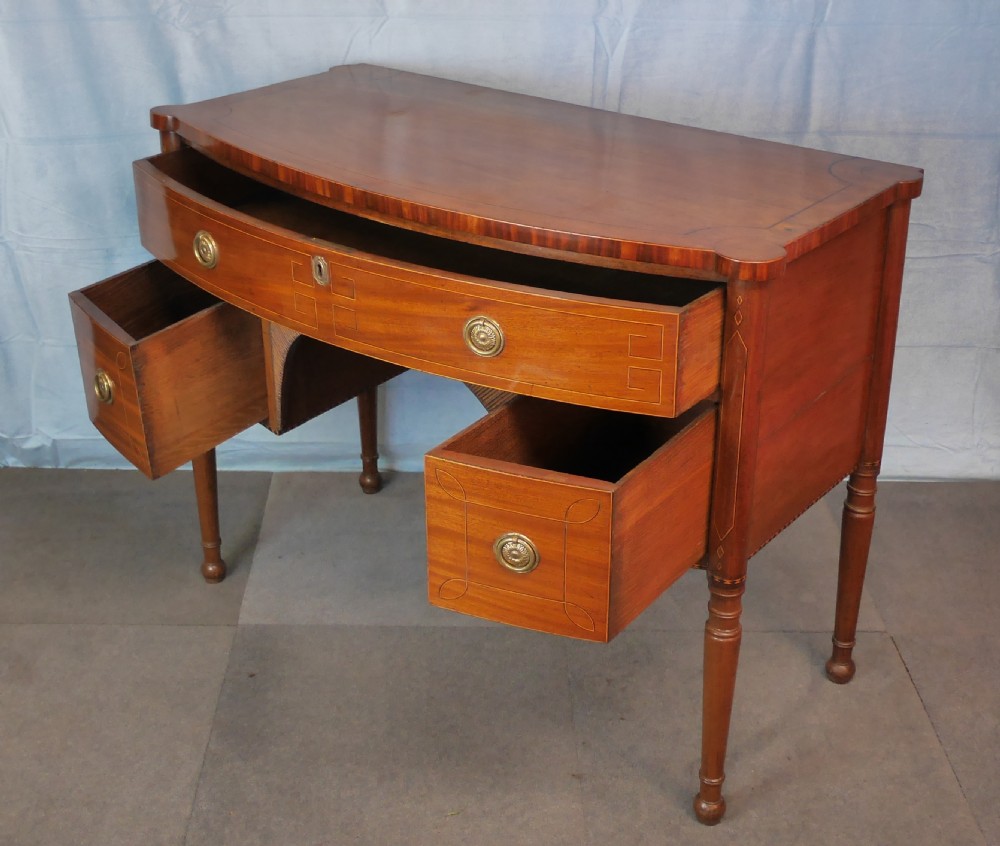 An antique Regency mahogany dressing table featuring a curved front, boxwood stringing inlay, three drawers with brass ring handles, and turned legs. View V