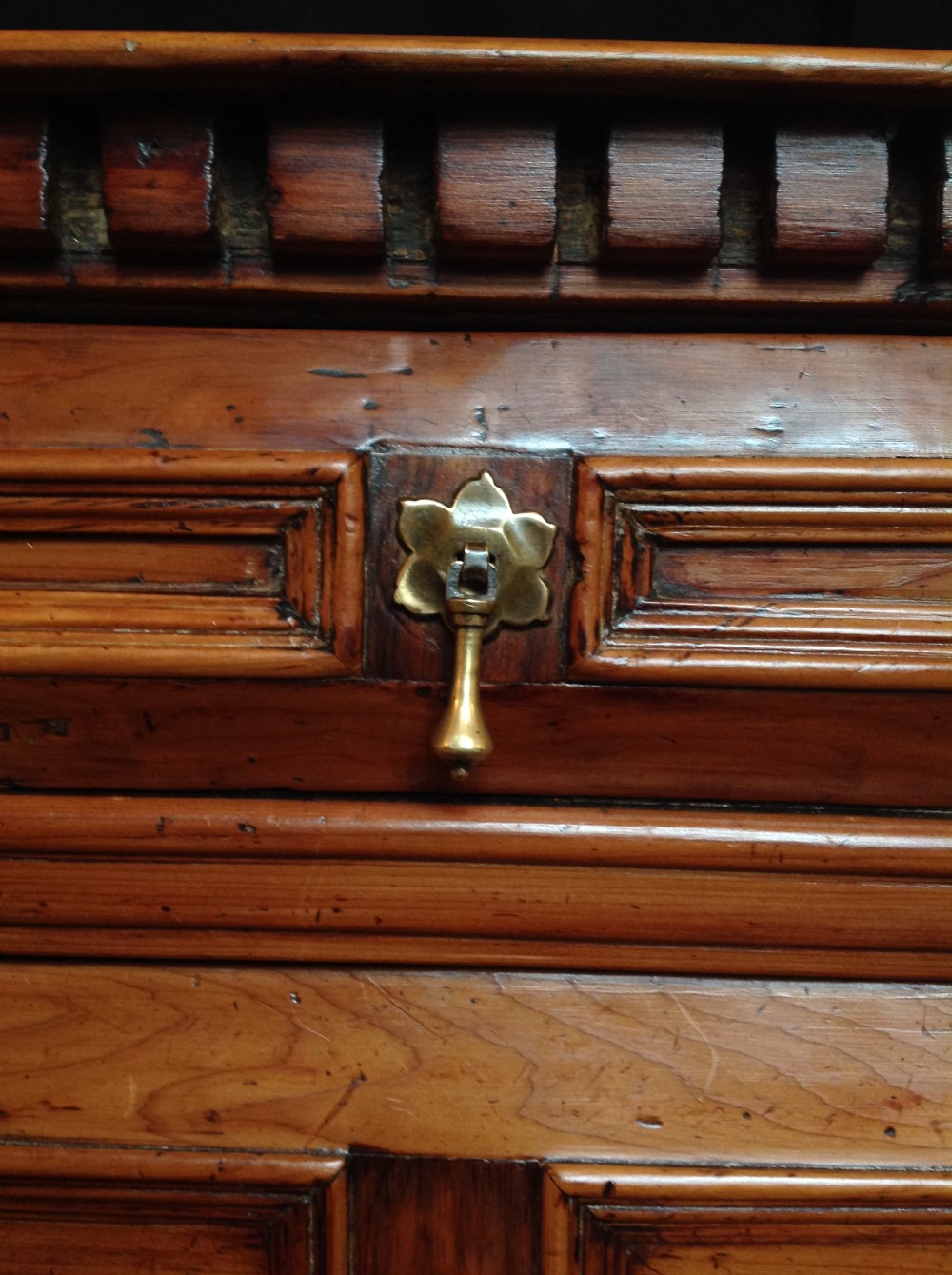 A seventeenth-century cedar chest of drawers featuring geometric moulded drawer fronts, brass drop handles, ornate escutcheons, and bun feet. View VII