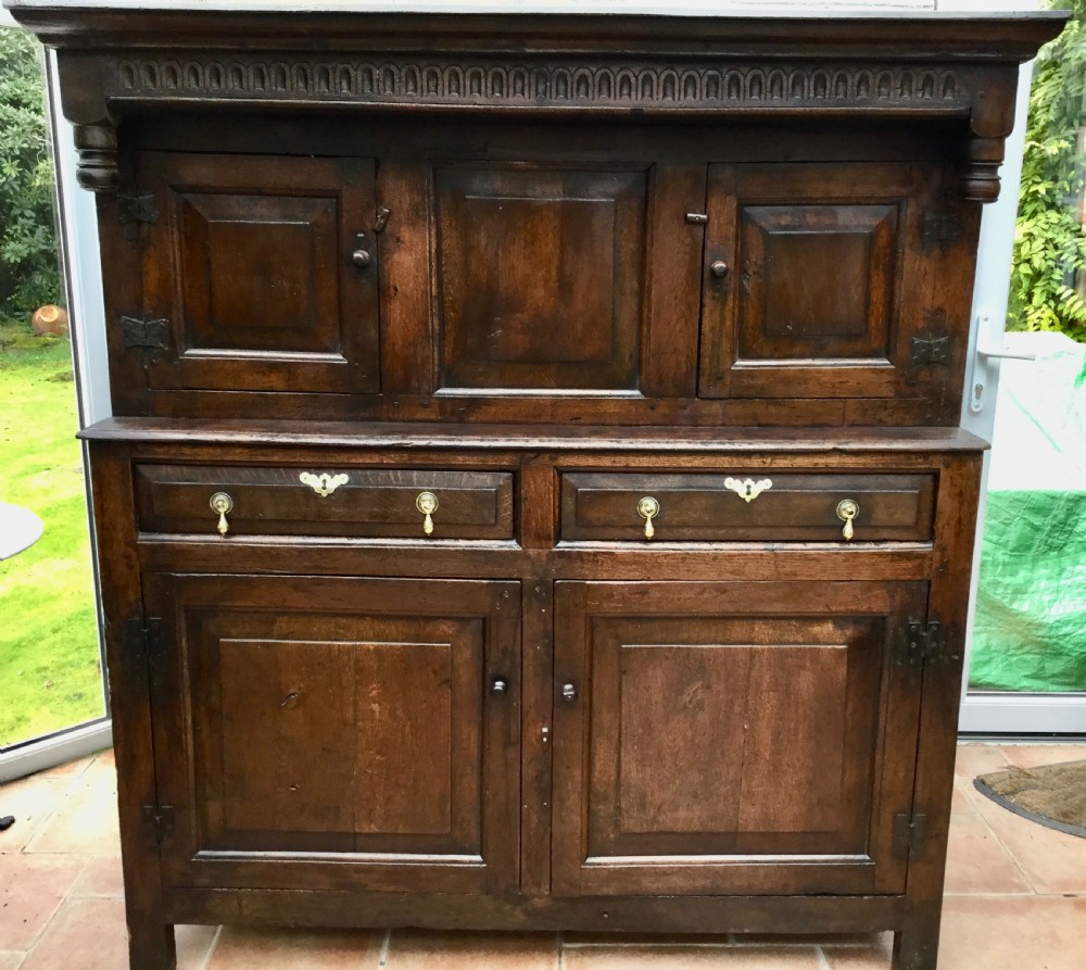 A dark oak 17th-century court cupboard with geometric fielded panelling, carved frieze, pendant drops, two drawers, and double lower doors.