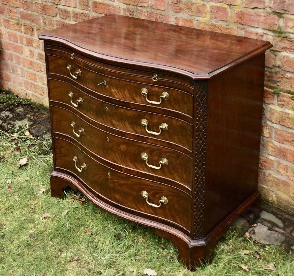 An antique dark mahogany serpentine chest of drawers featuring four graduated drawers, brass handles, and carved blind fretwork details on the corners.