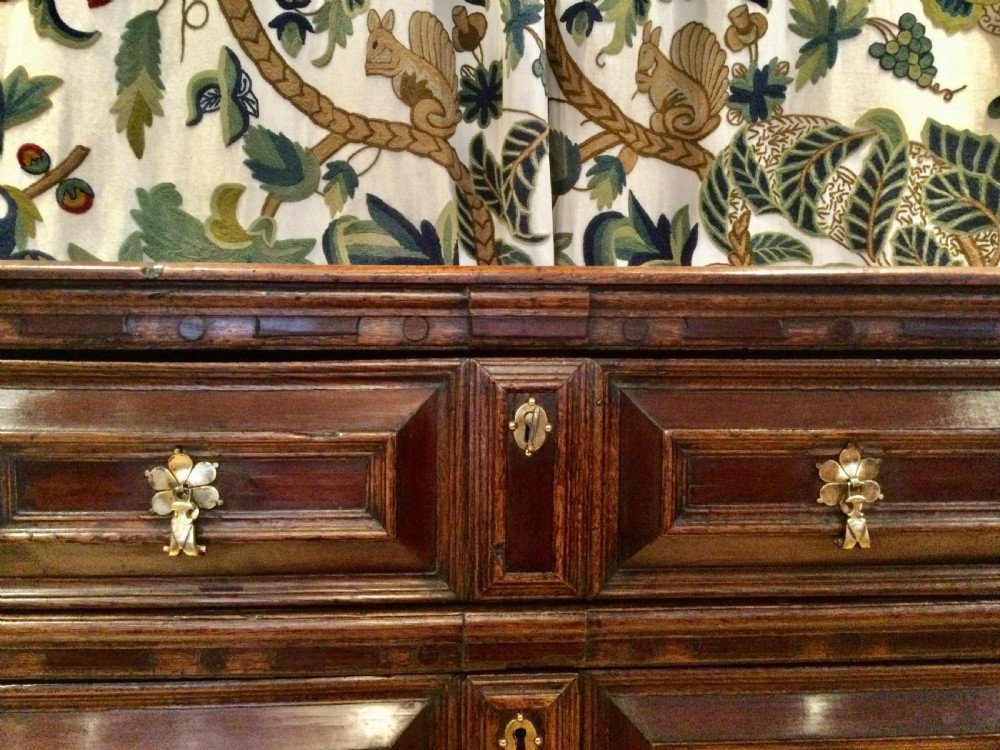 A dark brown seventeenth-century oak chest of drawers with geometric moulded panel fronts, brass drop handles, and bun feet. View VI