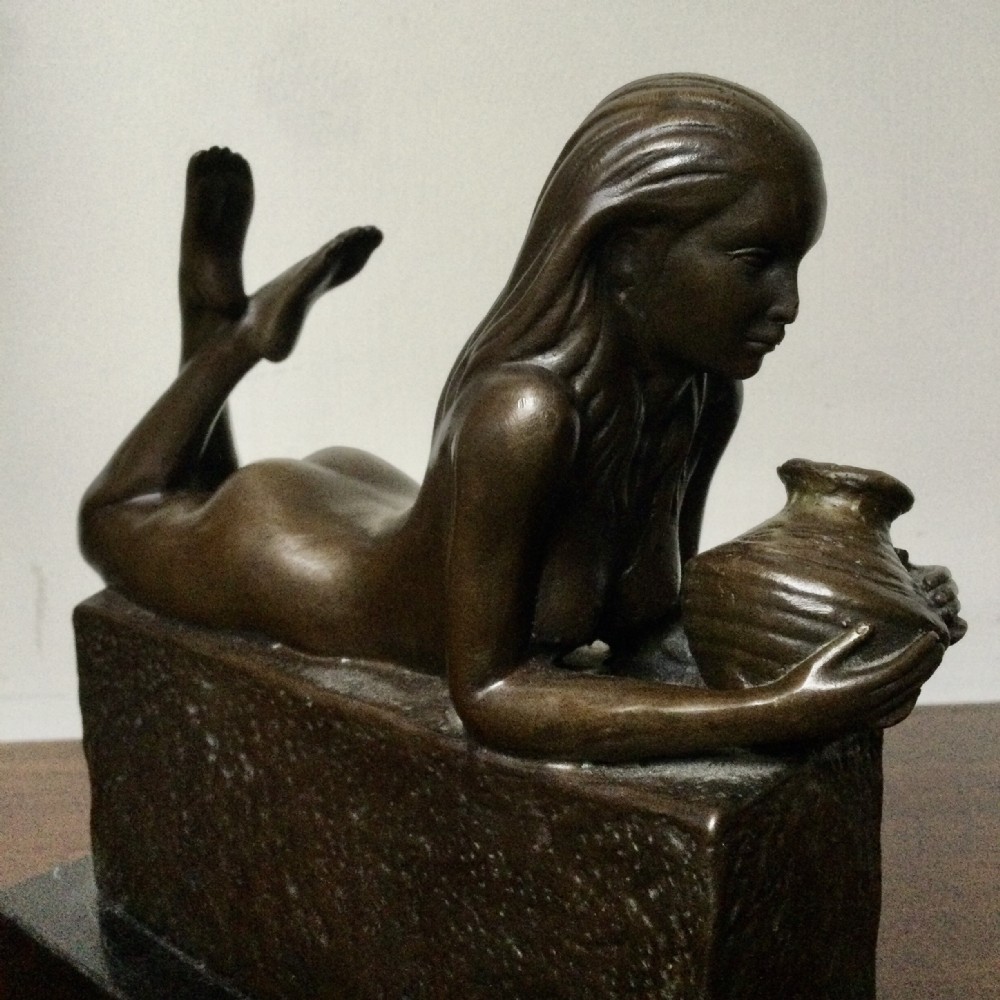 A dark patinated bronze sculpture depicting a reclining female nude figure holding a small vessel while resting on a rectangular base.