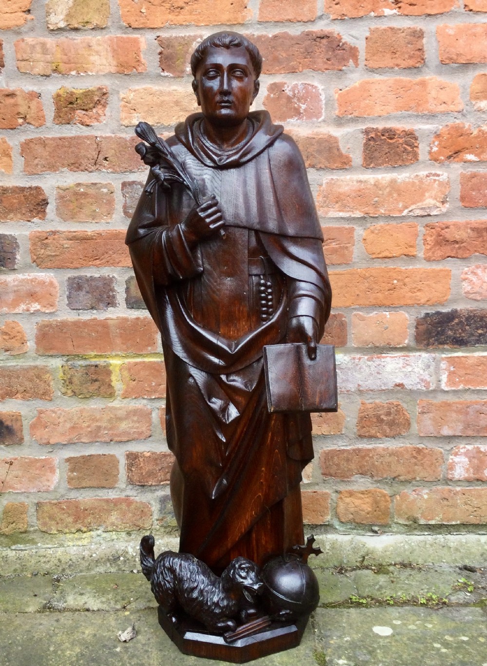 A dark carved oak sculpture of a religious figure holding a palm frond and open book with a carved animal at the base.