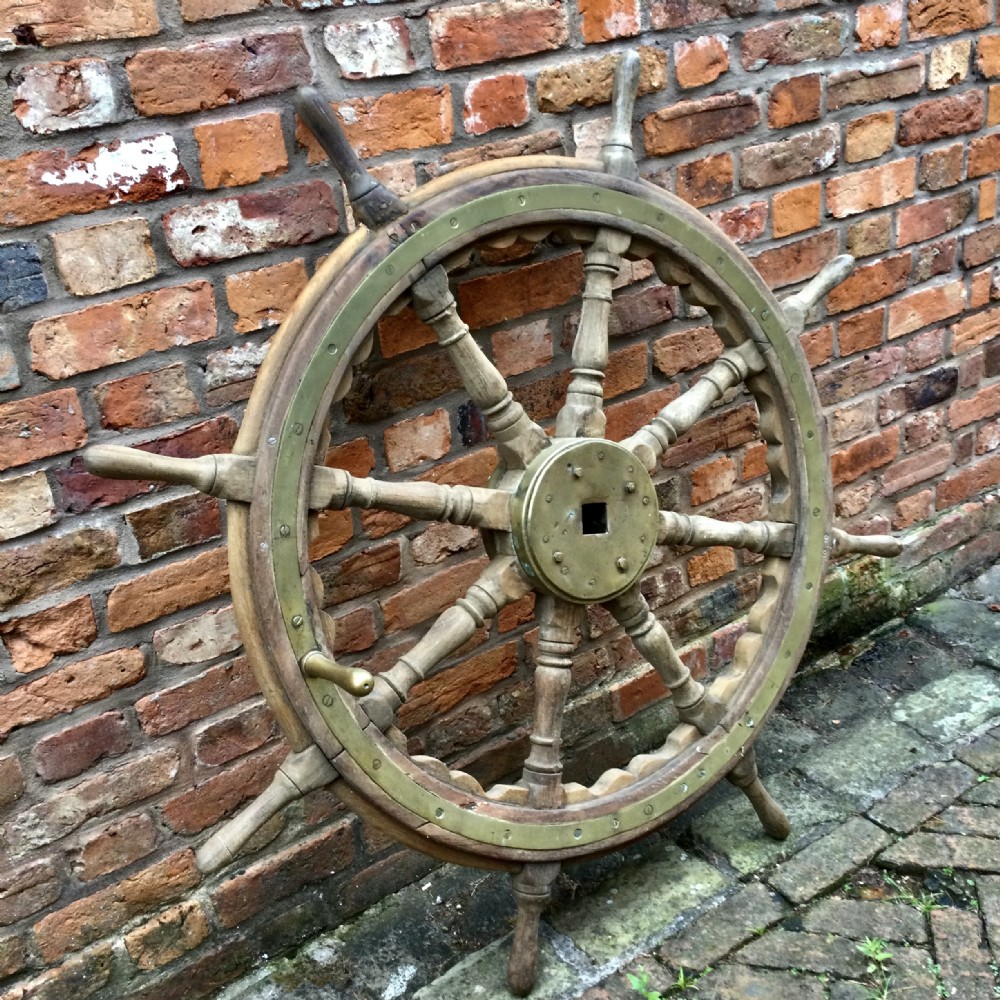An antique wooden ship helm featuring turned handles, a circular wooden rim reinforced with brass banding, and a central brass hub.