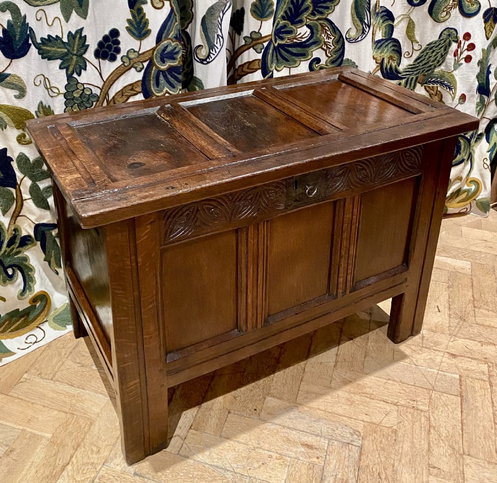 An antique dark oak coffer featuring three carved front panels, a hinged panelled lid, and raised stile legs.
