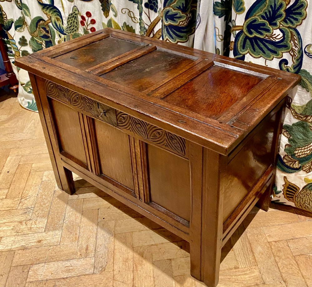 An antique dark oak coffer featuring three carved front panels, a hinged panelled lid, and raised stile legs. View III