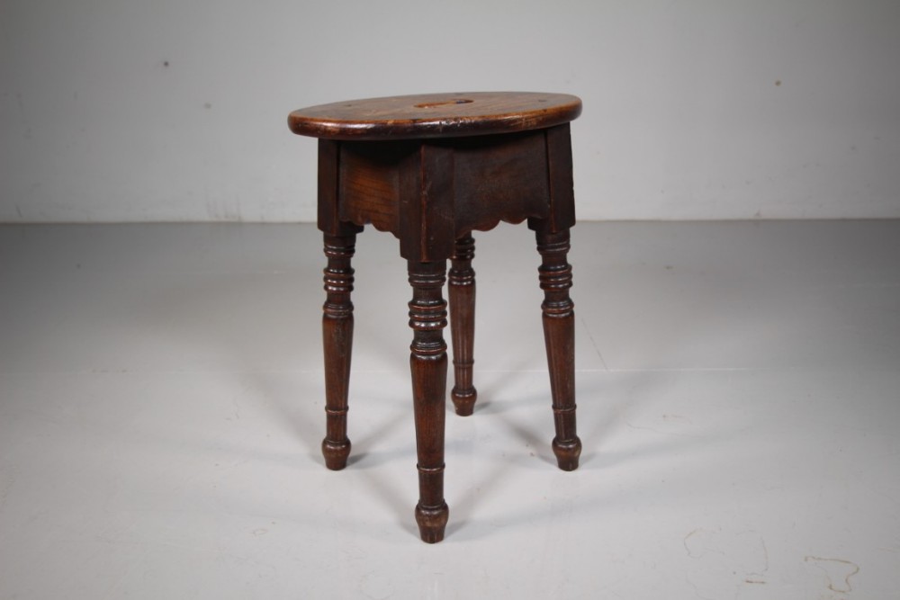 An antique dark brown elm stool featuring a round top, shaped apron, and four turned legs.