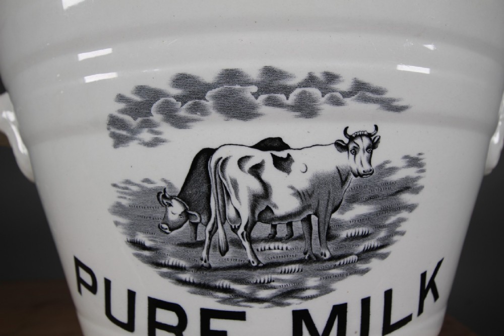 A white glazed ironstone dairy pail featuring integrated side handles, horizontal ribbed detailing, and a black transferware illustration of cows above the text PURE MILK. View II
