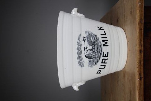 A white glazed ironstone dairy pail featuring integrated side handles, horizontal ribbed detailing, and a black transferware illustration of cows above the text PURE MILK. View VI