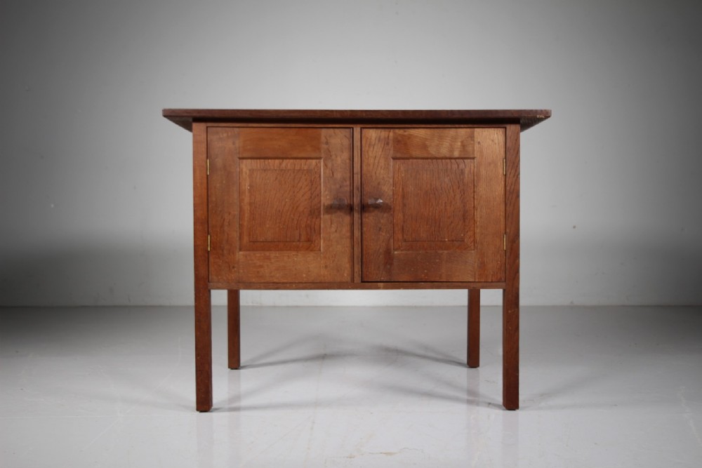 A solid oak two-door cupboard featuring recessed square door panels, carved wooden handles, and four square straight legs.