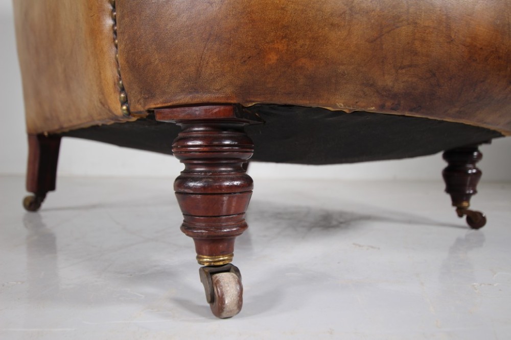 A 19th-century English leather armchair featuring deep-buttoned upholstery in tan brown hide, rolled arms, and turned mahogany front legs with brass castors. View IV