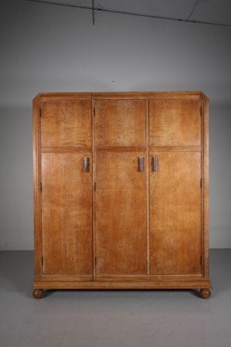 A 1920s Heals solid oak triple-door wardrobe with chamfered top corners, vertical wooden handles, and bun feet.