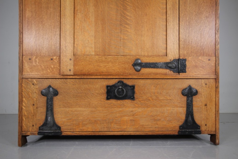 An Arts and Crafts solid oak cupboard featuring hand-forged wrought iron strap hinges, a single panelled door, and a lower drawer. View IV