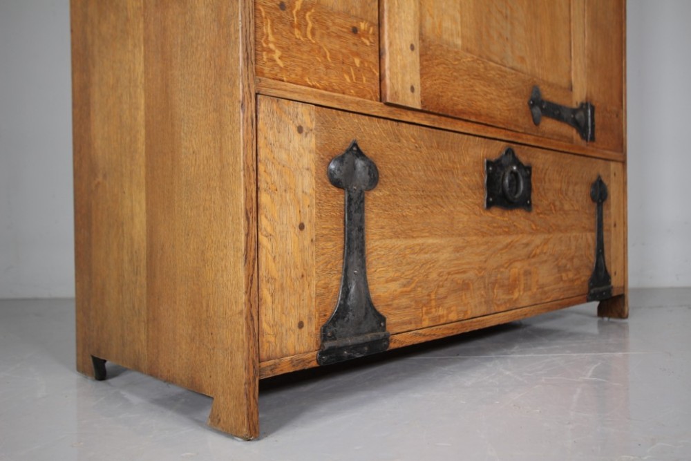 An Arts and Crafts solid oak cupboard featuring hand-forged wrought iron strap hinges, a single panelled door, and a lower drawer. View VIII