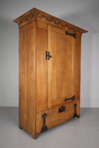 An Arts and Crafts solid oak cupboard featuring hand-forged wrought iron strap hinges, a single panelled door, and a lower drawer.