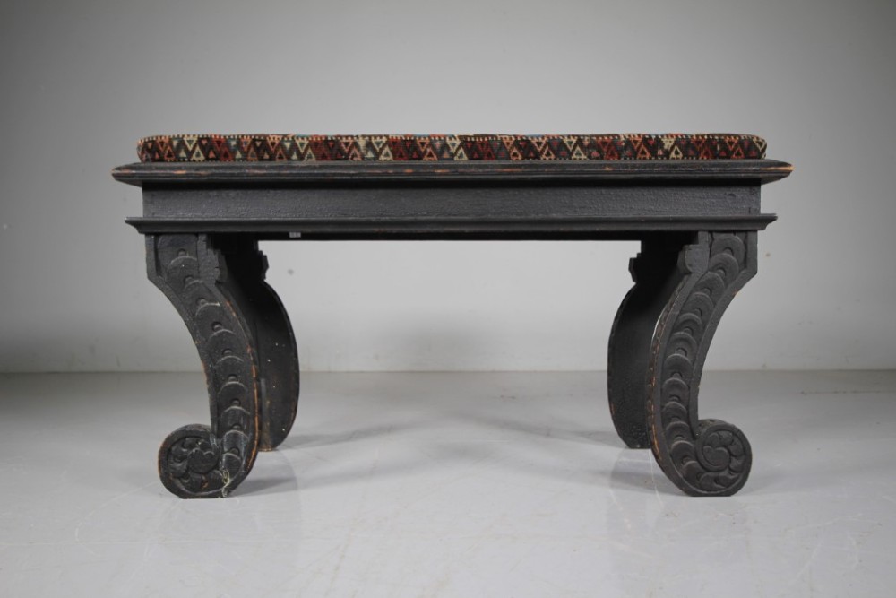 An early nineteenth-century black painted pine window seat featuring carved scroll legs decorated with repeating motifs and an upholstered geometric pad.