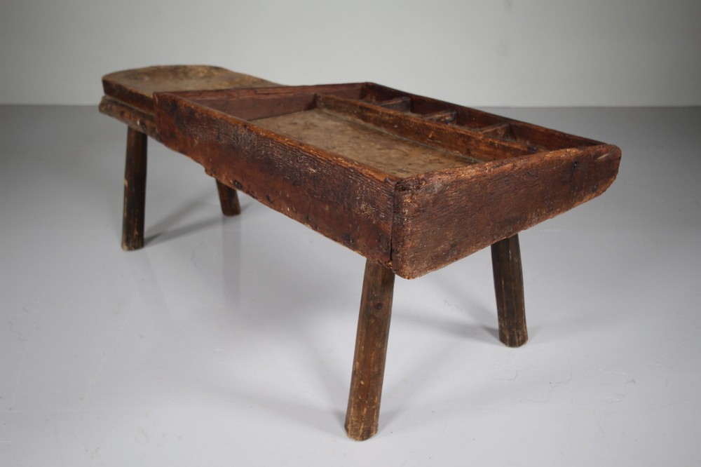 A 19th-century English wooden cobblers bench featuring a flat seating area on one side and a partitioned tray with tool compartments on splayed wooden legs. View V