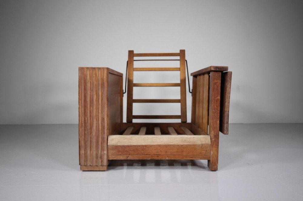 A 1930s solid oak reclining armchair frame featuring integrated side bookshelves, an adjustable slatted backrest, and a folding drop-leaf side table. View VII